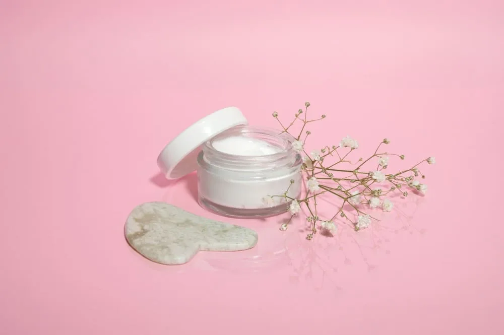 Pot de crème blanche ouvert avec accessoire en jade et fleurs sur fond rose pastel