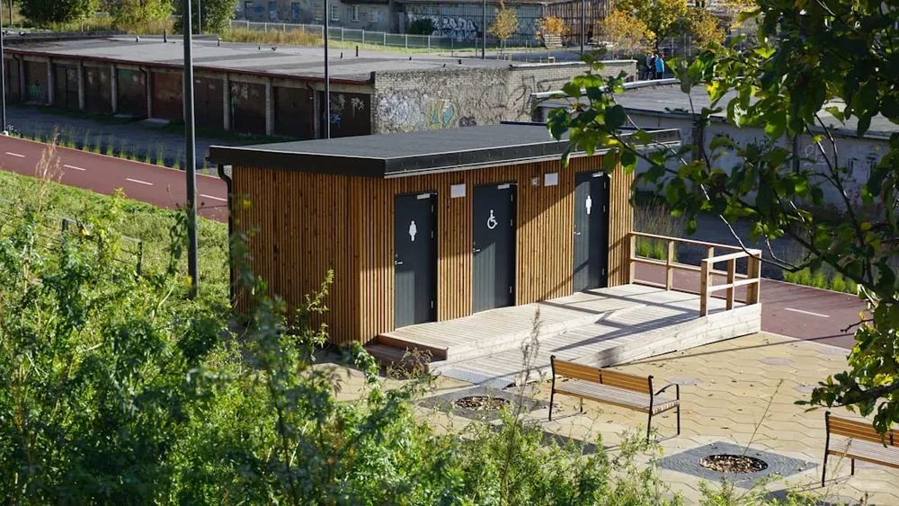 Trois portes de toilettes publiques en bois avec signalétique située dans un parc arboré