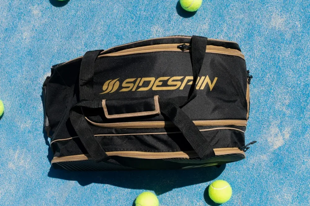 Sac de sport noir posé sur un court bleu entouré de balles de tennis.